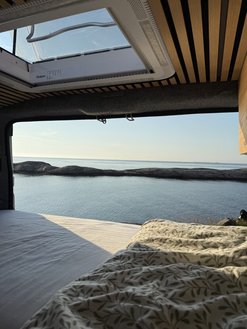 Blick aus dem Bett eines FeinWerk Campervans durch die geöffneten Hecktüren auf eine ruhige Meeresbucht am Abend